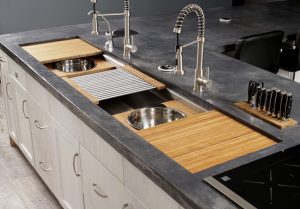 Graphite concrete counter top with two faucets over a galley kitchen sink and its cutting board and colander accessories.