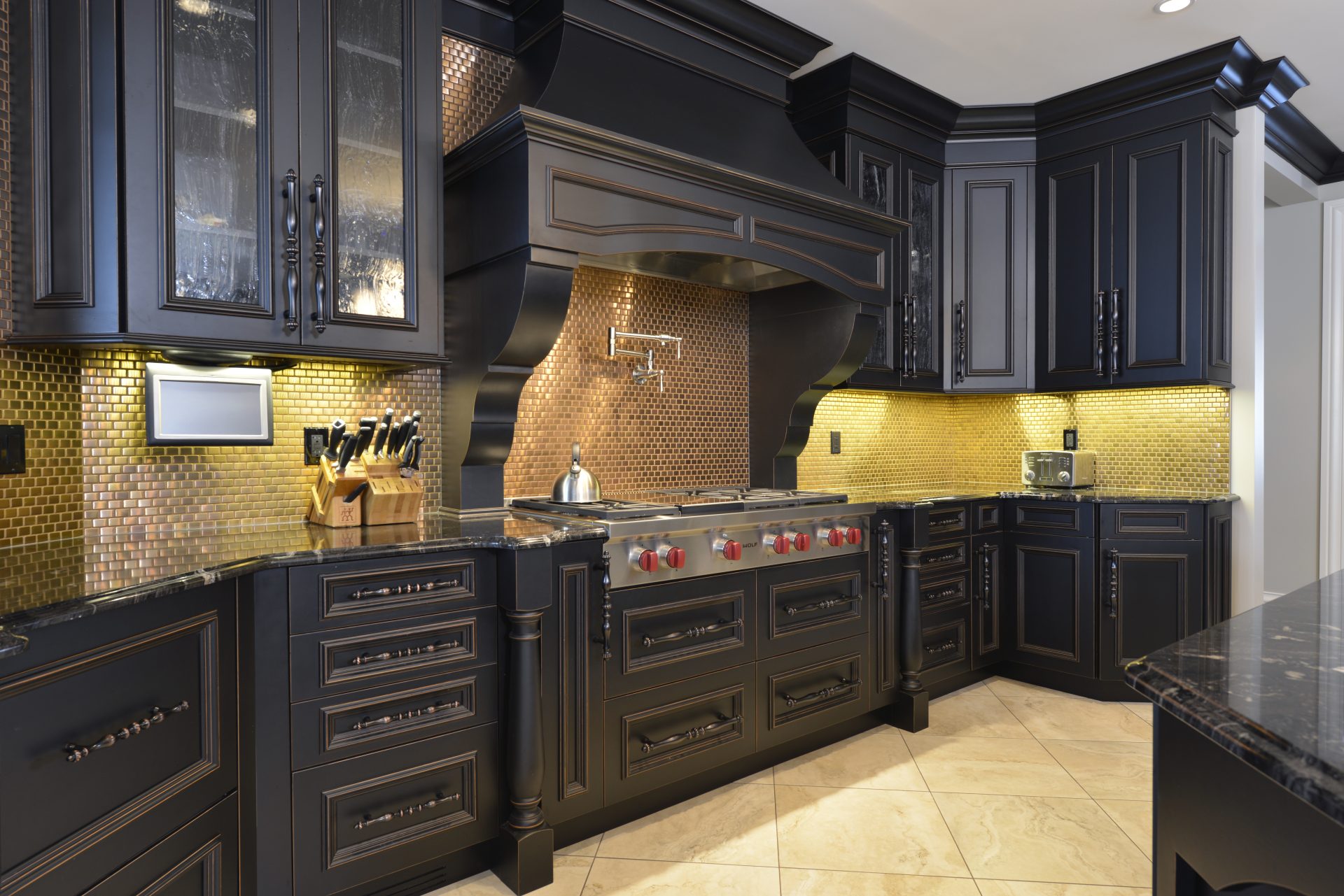 Dark cabinets in a kitchen with a bronze blacksplash and a silver pot filler over the stove