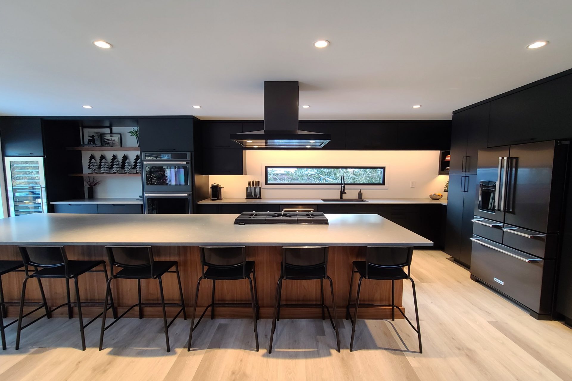 Modern kitchen featuring matte black cabinetry, large wood-accented island with seating, sleek appliances, and minimalist open shelving under recessed lighting.