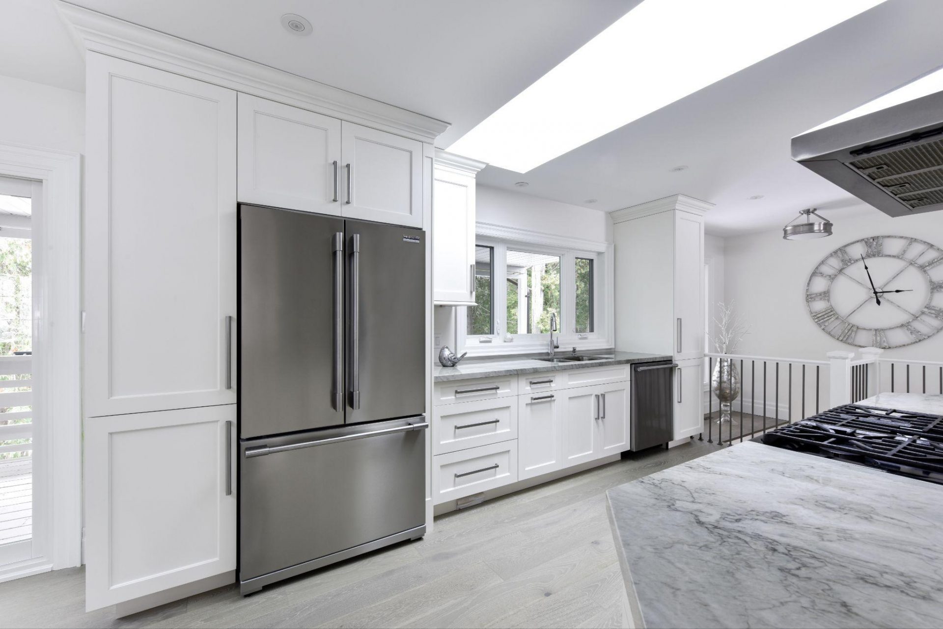 A bright kitchen with a large stainless steel refrigerator, white cabinets, and a spacious countertop, accented by a large wall clock and natural light from overhead skylights.