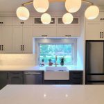 Modern two-tone kitchen with white upper cabinets, dark lower cabinets, farmhouse sink under a large window, stainless steel refrigerator, and a gold chandelier with globe lights.