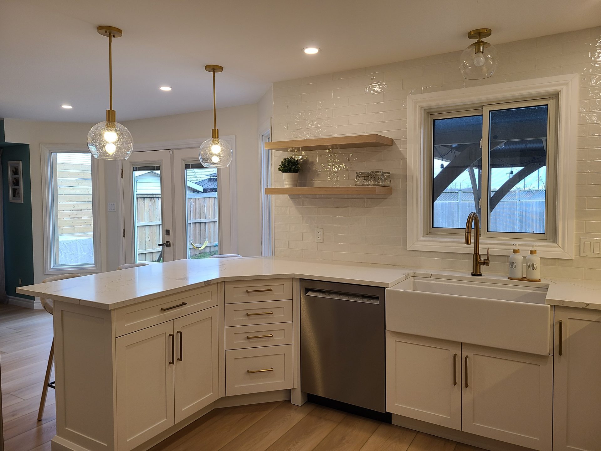Bright modern kitchen with white shaker cabinets, farmhouse sink, gold hardware, marble countertops, open wood shelves, and globe pendant lights.