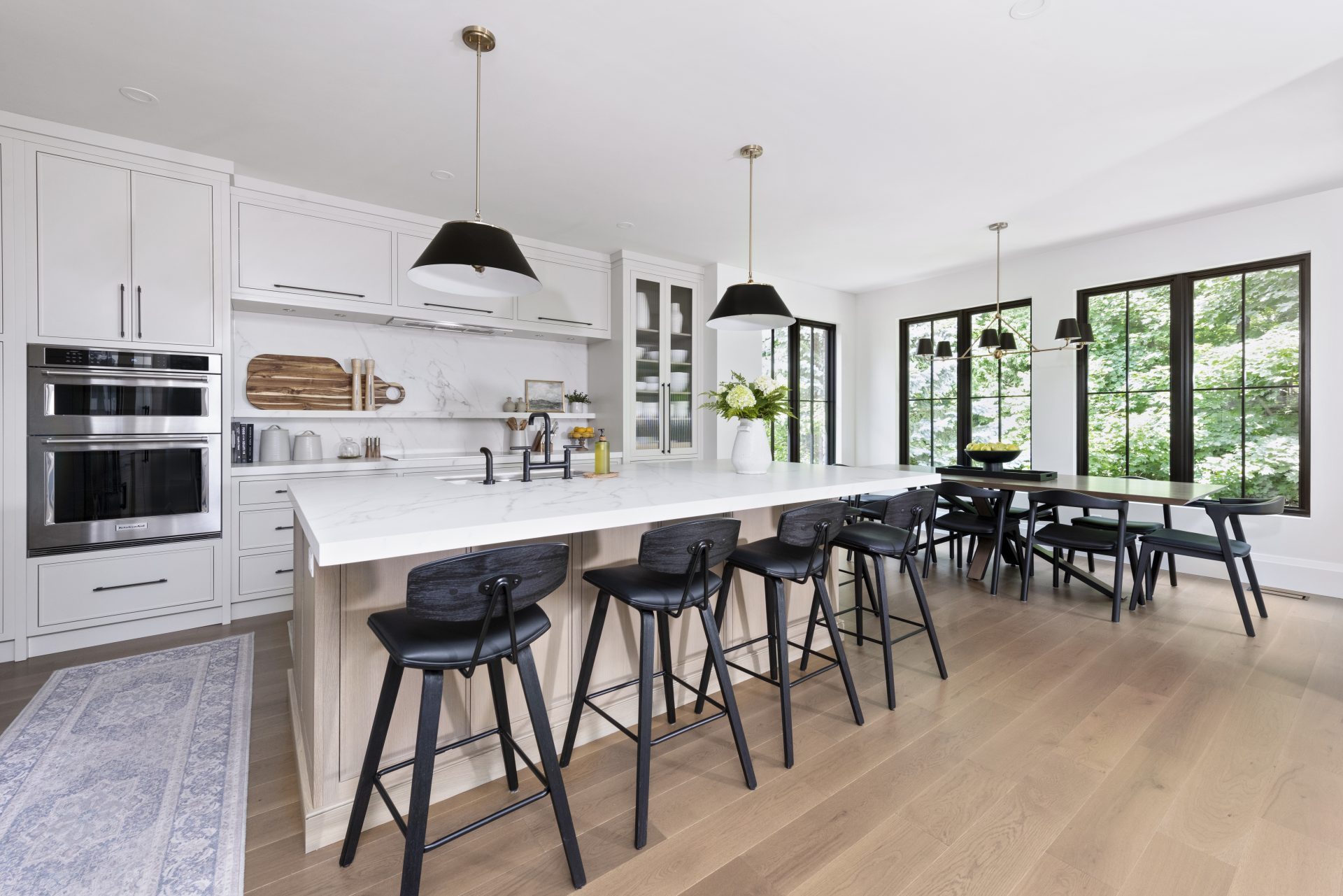 Contemporary kitchen with wood grain cabinets, white quartz island with bar stools, modern black accents, stainless steel appliances, and a dining table with black chairs.