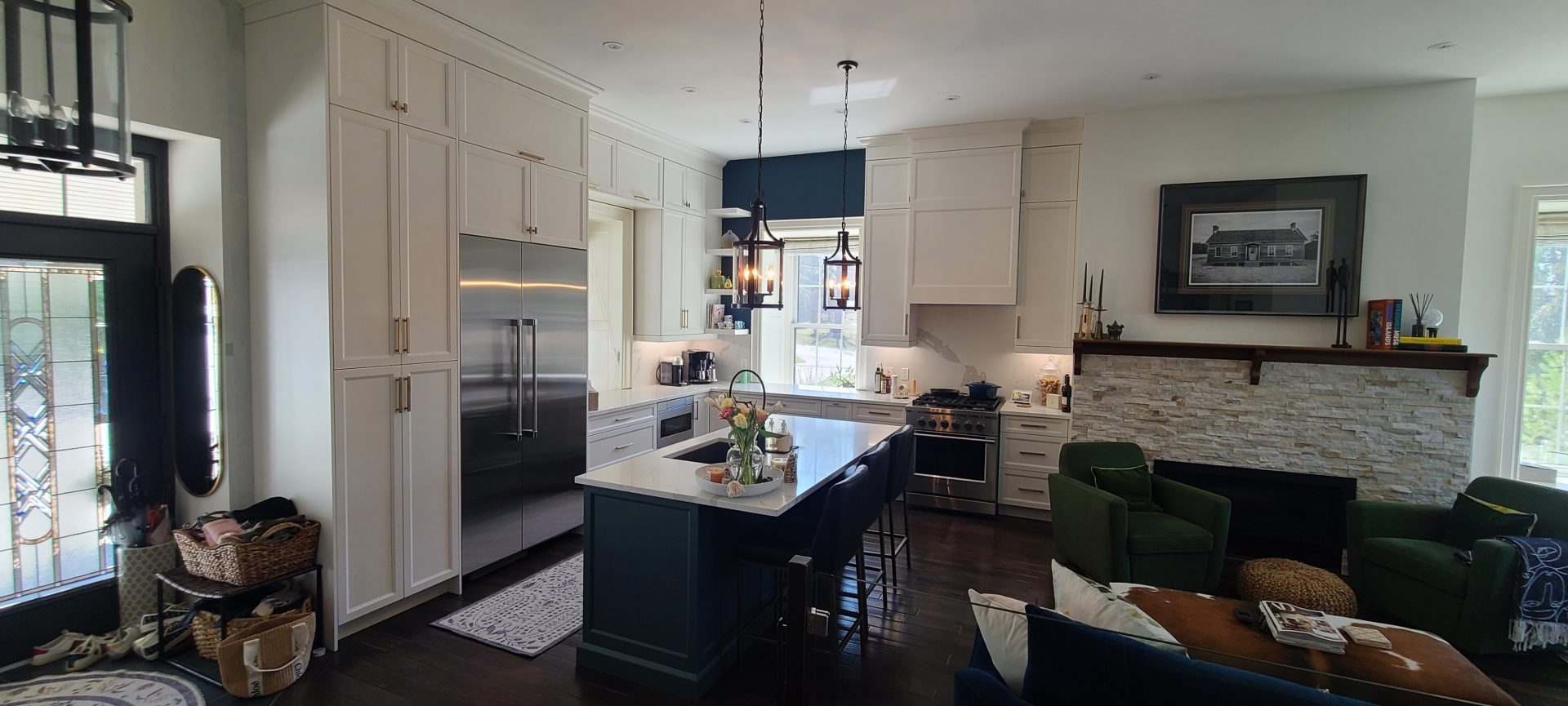 Open-concept white kitchen with navy blue island, brass lantern pendant lights, and a stone fireplace in the adjoining living area.