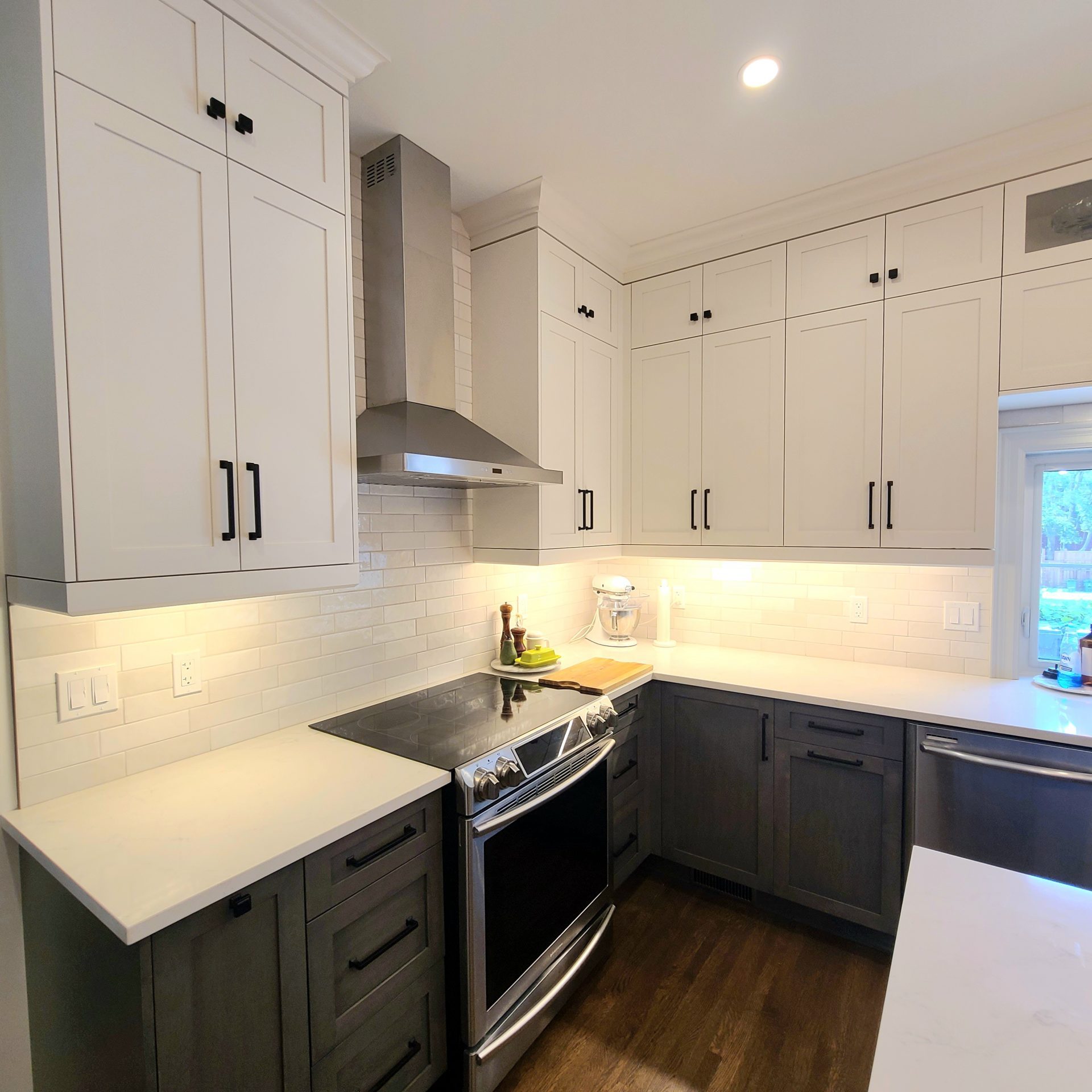 White upper cabinets with black hardware, stainless range hood, subway tile backsplash, and dark lower cabinets.