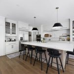 Bright white kitchen with a large marble-look island, black pendant lights, black bar stools, and glass-front cabinets.